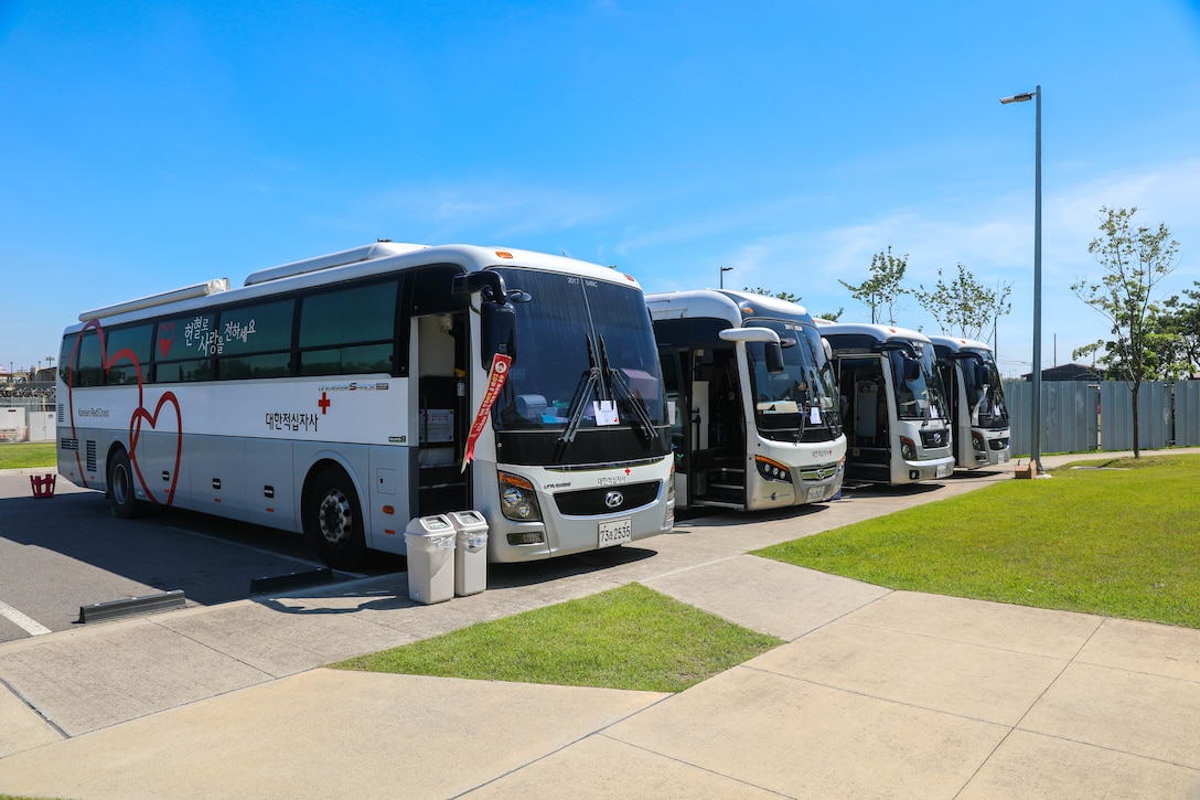 Four large KNRC donation buses are parked in a line at the blood donation location's parking lot. The sky is bright blue and it is very sunny.