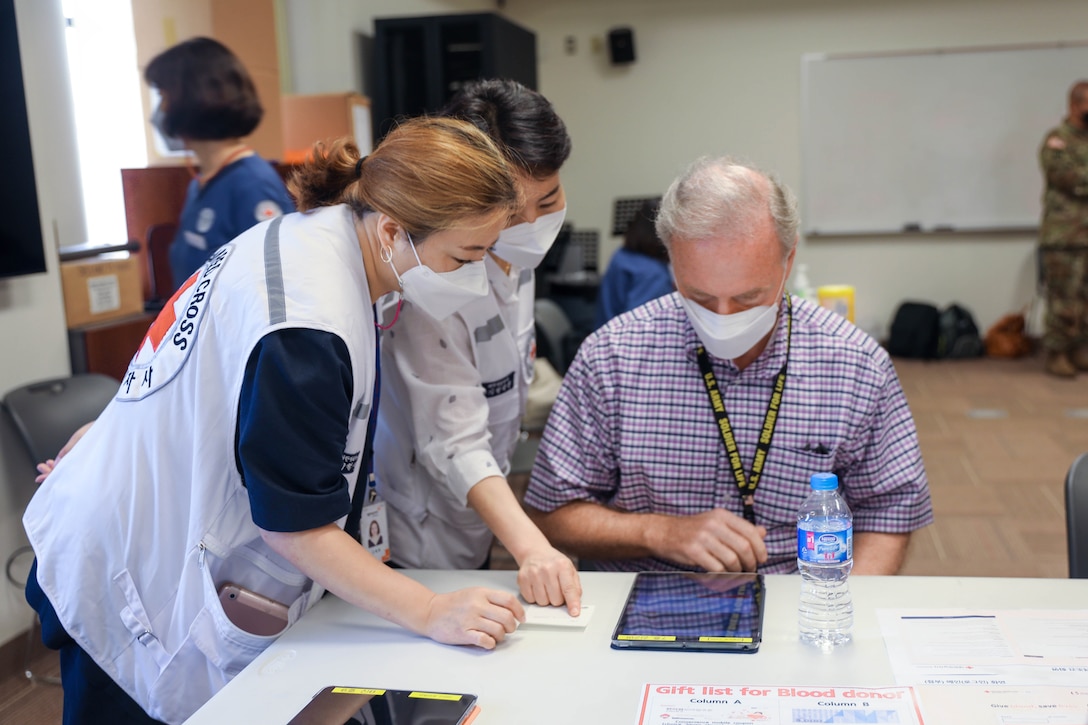 Members of the Korean National Red Cross, stand over a table pointing and assisting an older gentleman, who is sitting at the table, with answering questions in relations to blood drive screening.