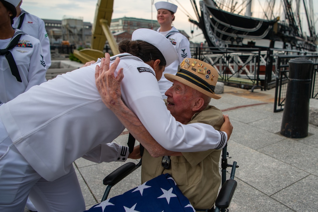 A sailor embraces an older man sitting with an American flag on his lap.