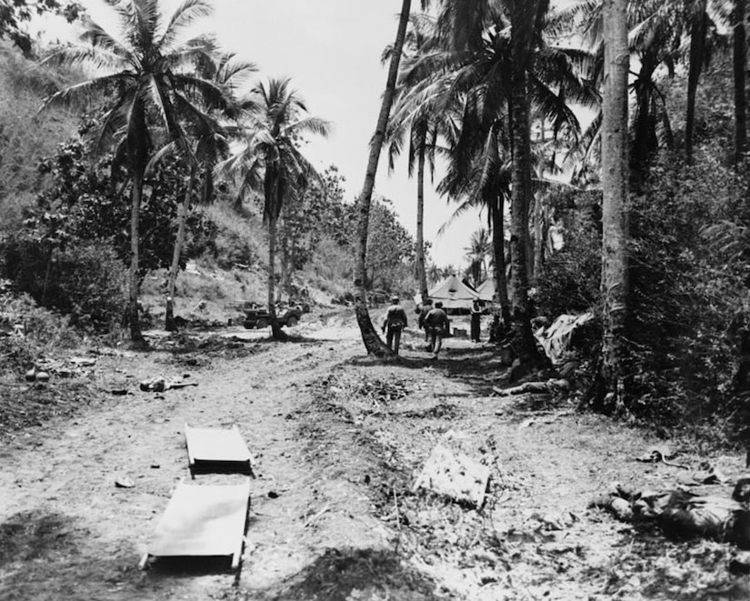 Two stretchers sit in a dirt roadway on a tropical island. A few men can be seen in the background.