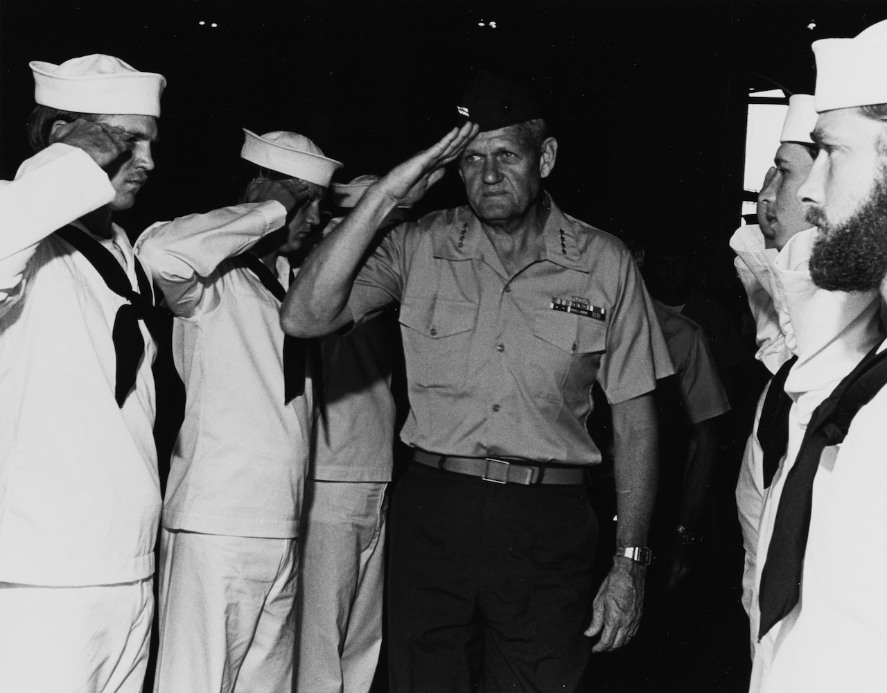 A man in a Marine uniform salutes sailors in dress white uniforms.