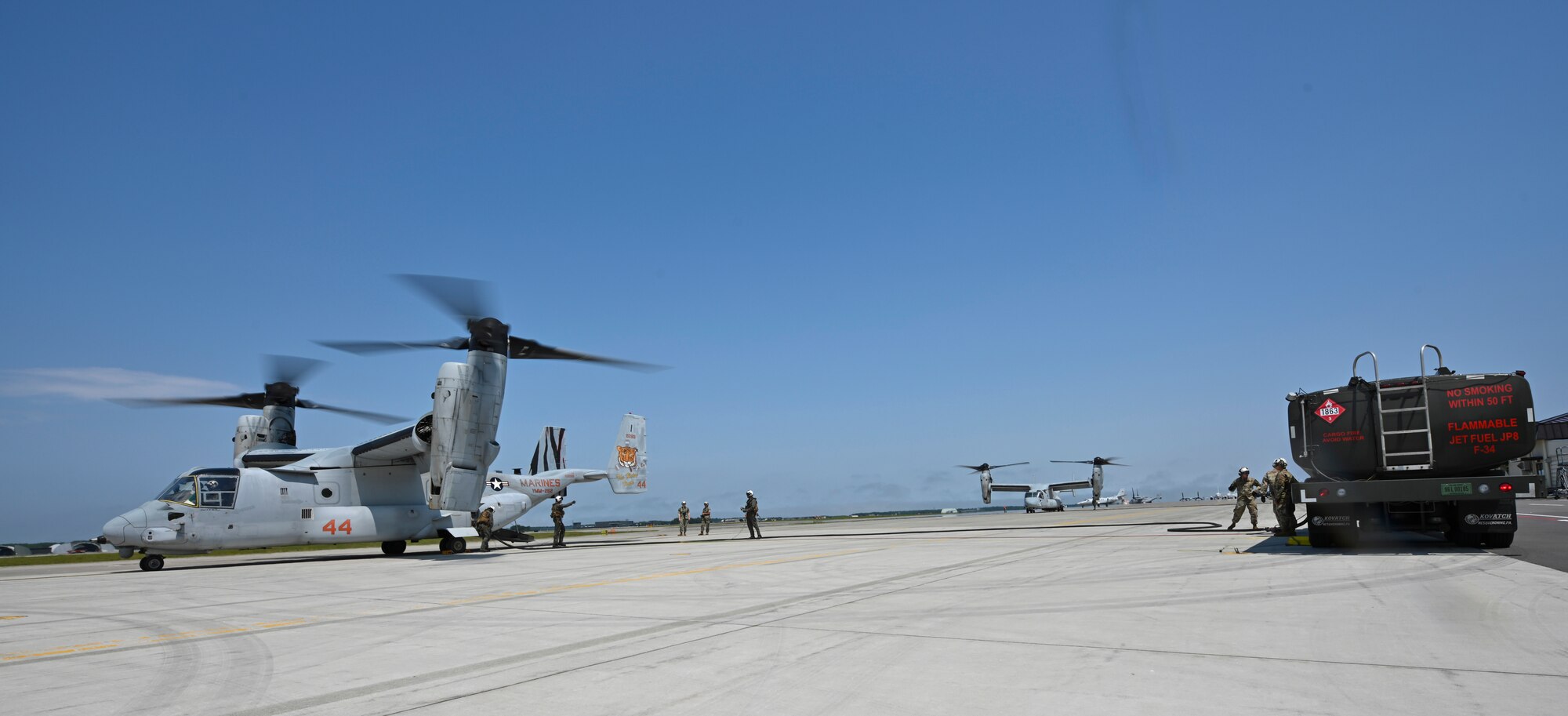 Service members stand next to a fuel truck separated from two tiltrotor aircraft.