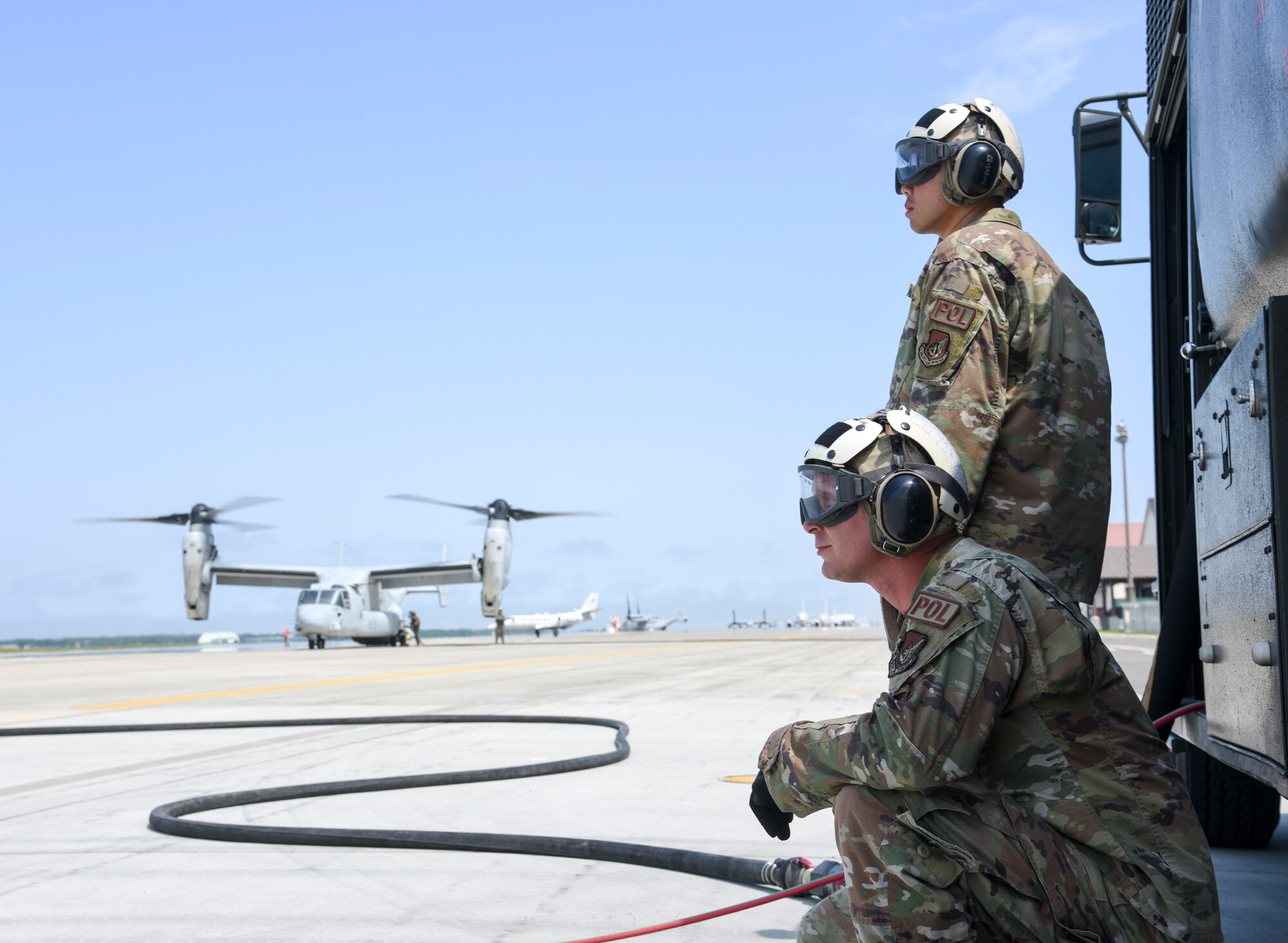 Two Airmen wait next to a truck while a tiltrotor aircraft is parked in the background.