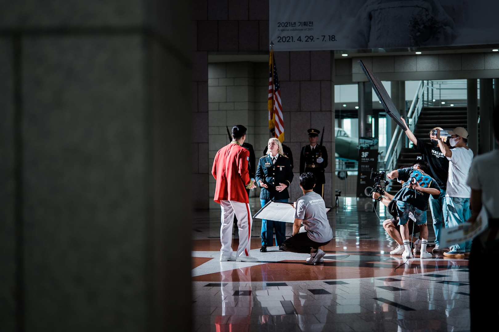 ROK Army Sgt. Yook Sung-jae in dress uniform and U.S. Army Spc. Brittany D. Simmons in dress uniform are standing in the center look at one another, while a video crew with cameras, lighting and sound are to the right of the frame. The left of the frame is blocked by a blurred wall. They're standing in the center of a rotunda located at the War Memorial of Korea.