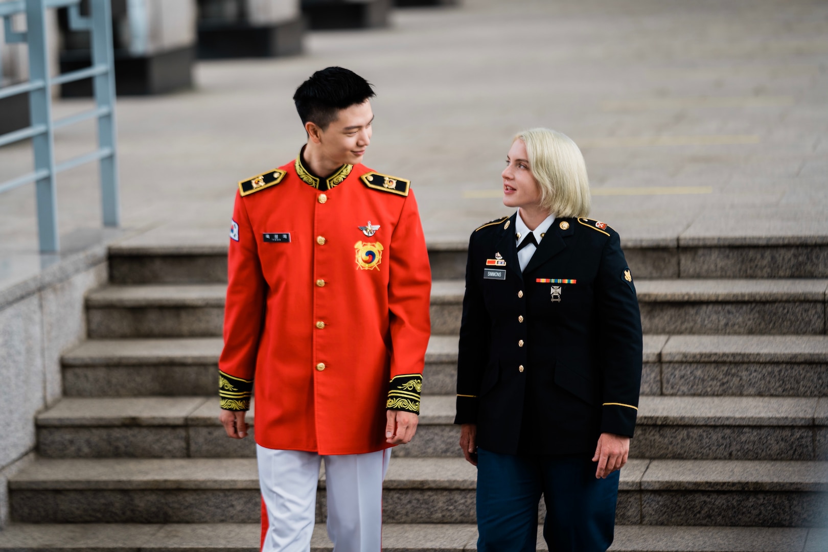 ROK Army Sgt. Yook Sung-jae in dress uniform is standing on the left while U.S. Army Spc. Brittany D. Simmons in dress uniform is standing on the right and both are looking at one another, talking. Behind them is a blurred stairway from inside of the War Memorial of Korea.