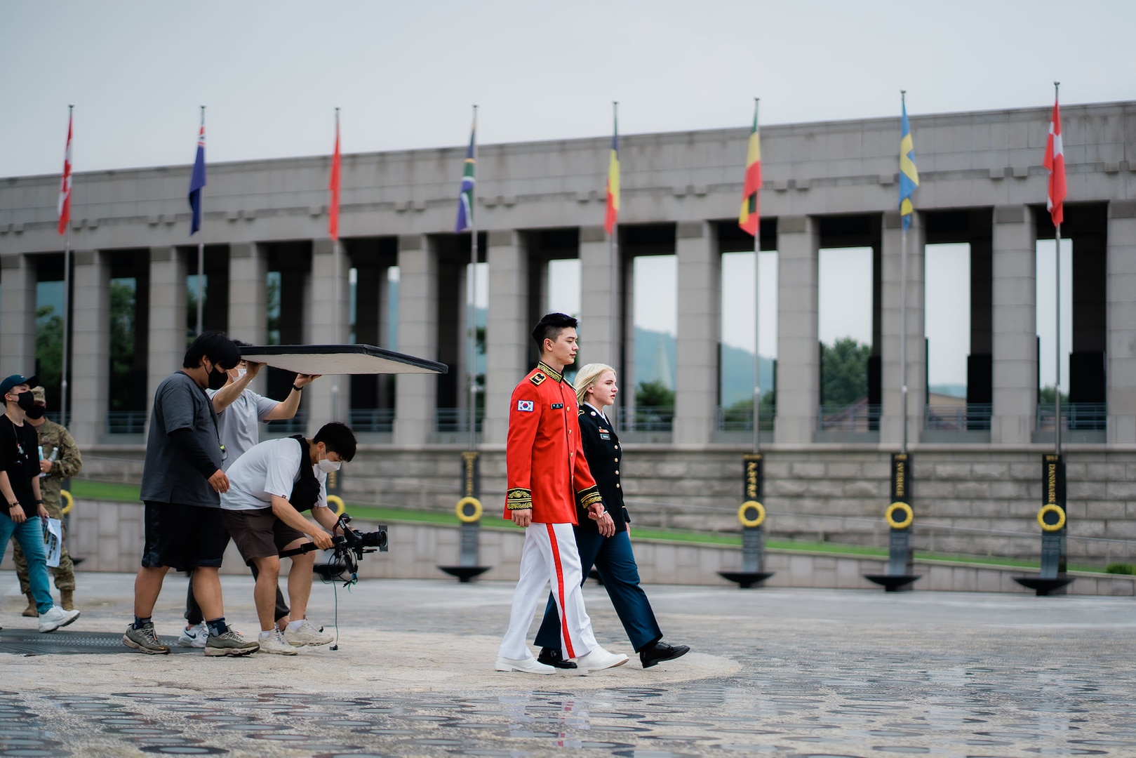 ROK Army Sgt. Yook Sung-jae in dress uniform and U.S. Army Spc. Brittany D. Simmons also in dress uniform are walking in the center with a video crew following behind. They are walking outside at the War Memorial of Korea in Seoul and there is a row of world flags behind them.