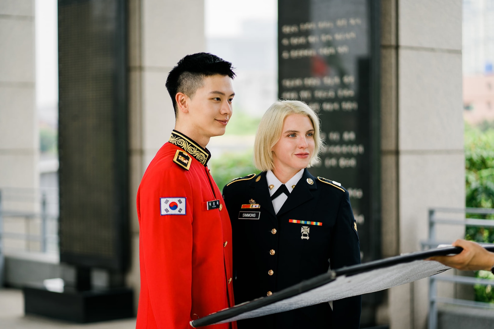 ROK Army Sgt. Yook Sung-jae stands in dress uniform next to U.S. Army Spc. Brittany D. Simmons in the center frame and looking off to the right. A person off screen holds a square photo reflector at the bottom. The background is at the War Memorial of Korea but blurred in the photo.