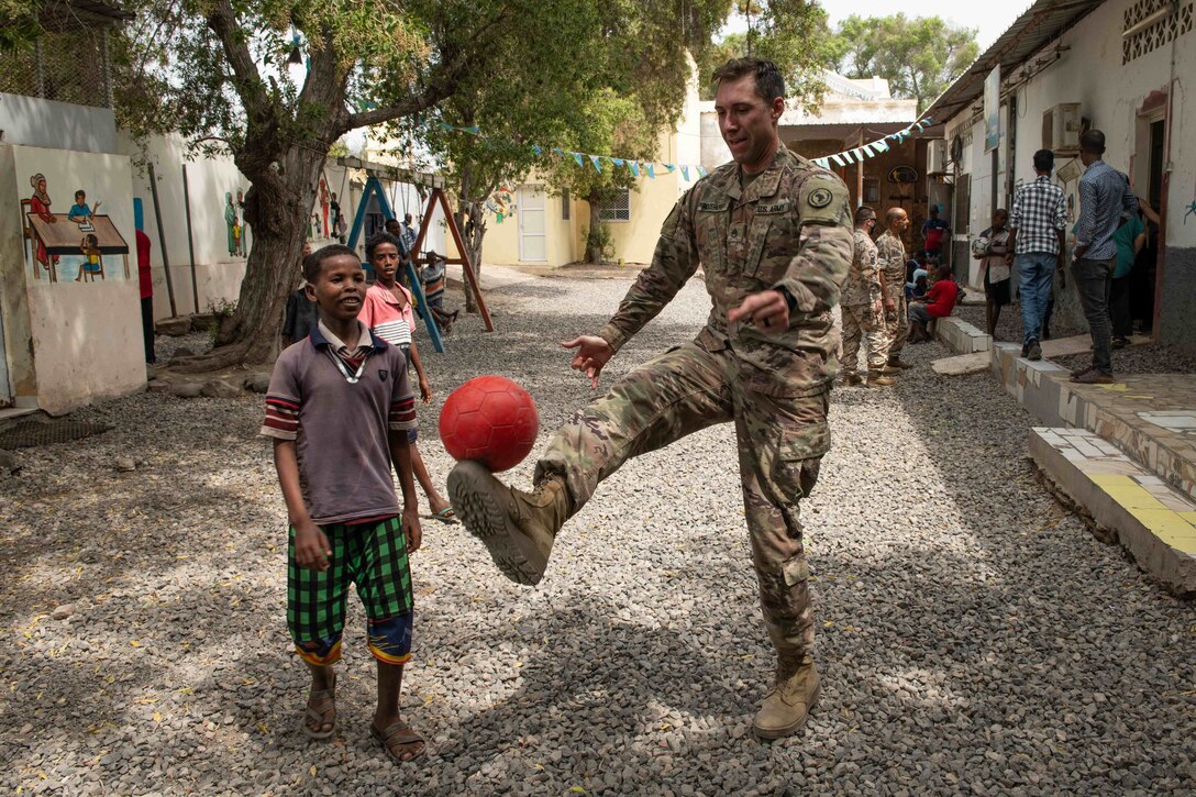 A soldier kicks a ball while a child watches.