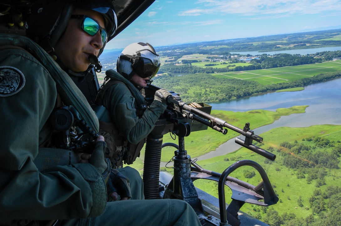 A Marine seated next to another on an airborne helicopter fires a machine gun.