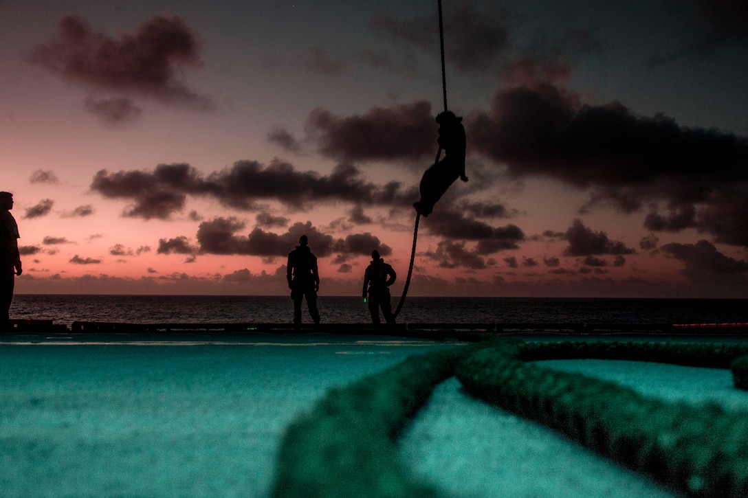 A Marine, shown in silhouette, maneuvers on a rope above a ship's deck as other Marines watch.