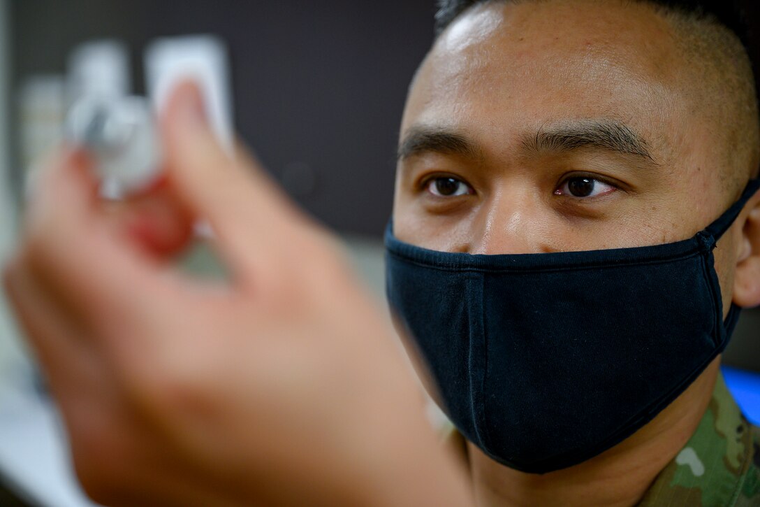 An airman wearing a facemask looks at a vial he's holding.