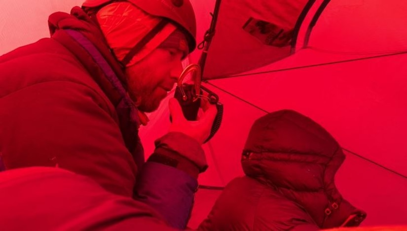 Two men clad in cold-weather great sit in a tent, one speaking into a radio.