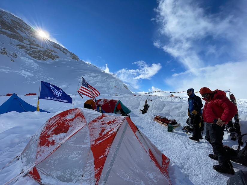 Two men stand near three snow-covered tents on the side of a snow-covered mountain.