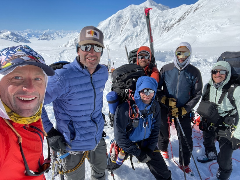 Six men clad in heavy winter gear pose for a photo on a snowy mountainside.