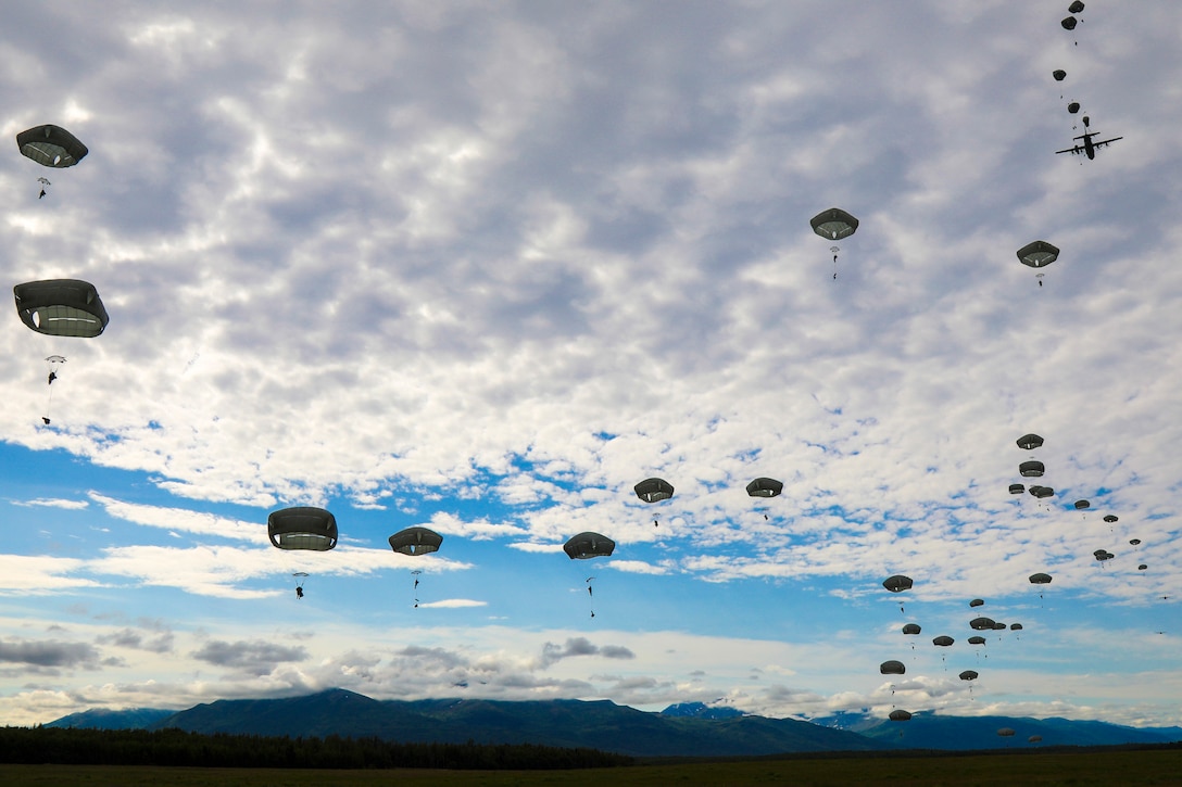 Soldiers wearing parachutes descend in the sky as a plane flies ahead.