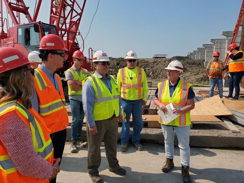 A group of individuals visit a construction site. There is a crane and cement wall behind the individuals.