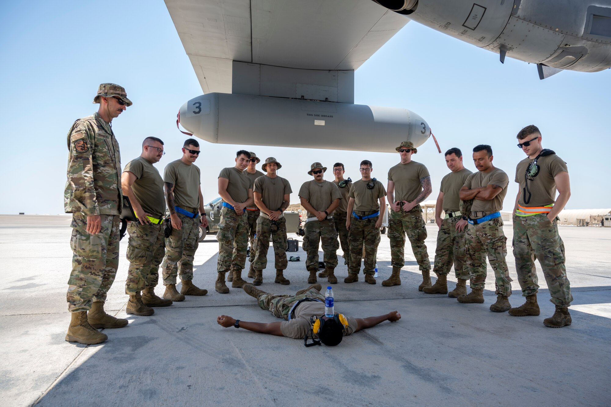 Airmen standing around one another Airman, who is lying on the ground with a water bottle on his head.