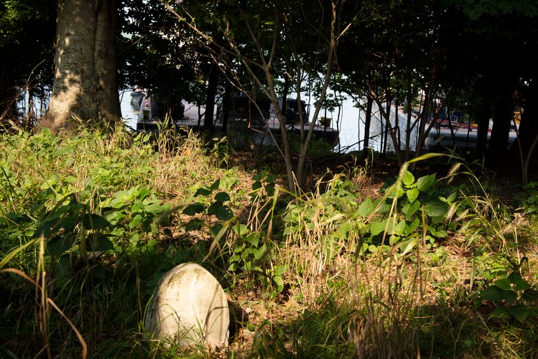 A headstone glimmers in the early morning sunlight as maintenance barges are positioned to install riprap on the shoreline of Old Hickory Lake July 22, 2021 to stop erosion from inching closer to the historic Rock Castle Slave Cemetery in Hendersonville, Tennessee. The U.S. Army Corps of Engineers Nashville District's Maintenance Support Team reshaped the embankment and laid down riprap July 19-22, 2021 where water had been undercutting the shoreline. (USACE Photo by Lee Roberts)