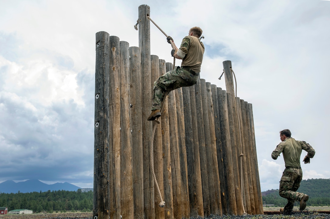 A soldier climbs a log fence using a rope.