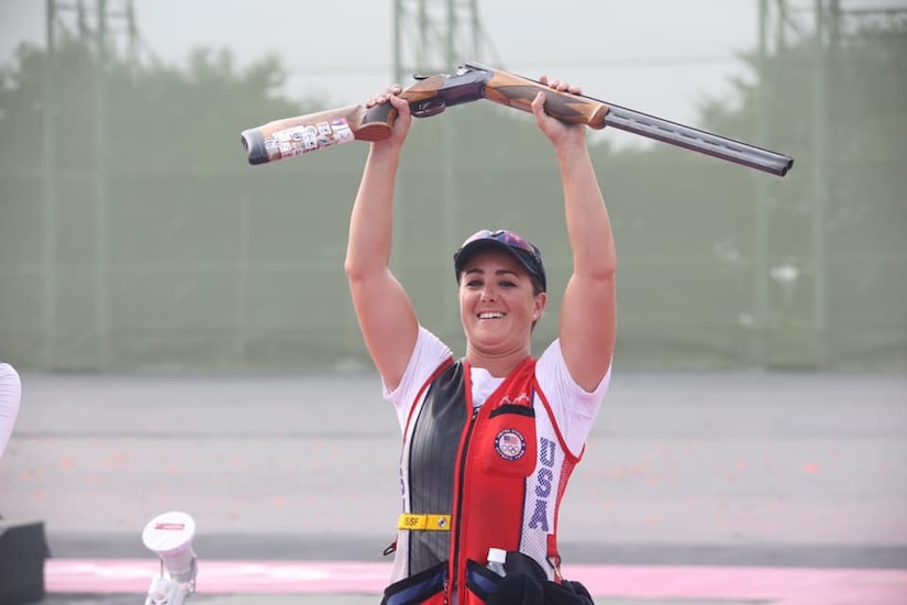 An athlete smiles as she holds a rifle above her head.