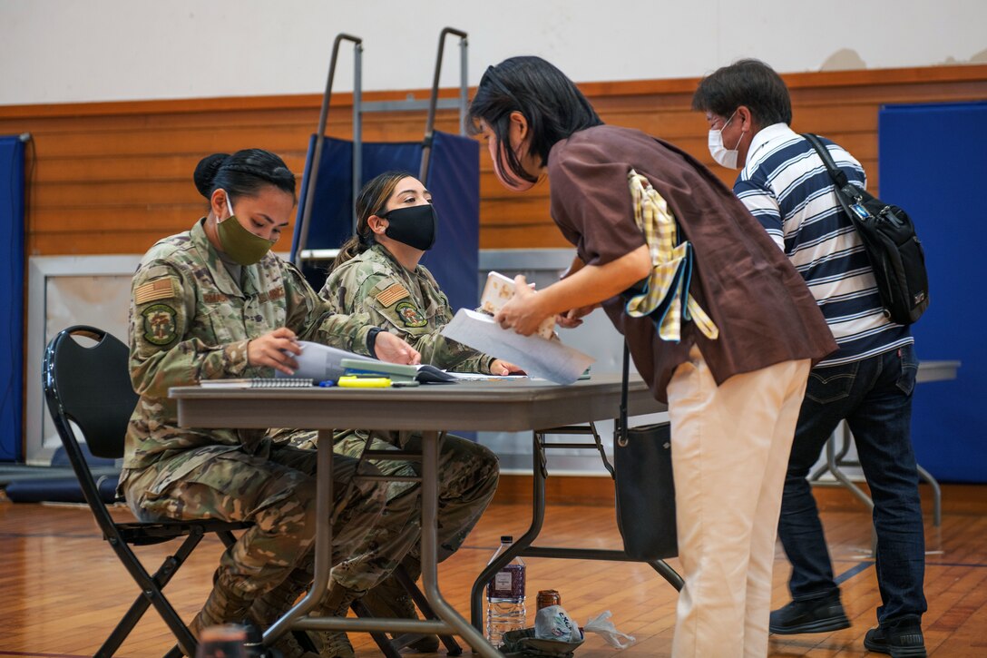 Two soldiers wearing face masks sit at a table and talk to two people, who are also wearing face masks.