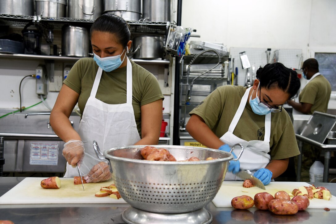 Two soldiers wearing face masks cut potatoes in a large kitchen.