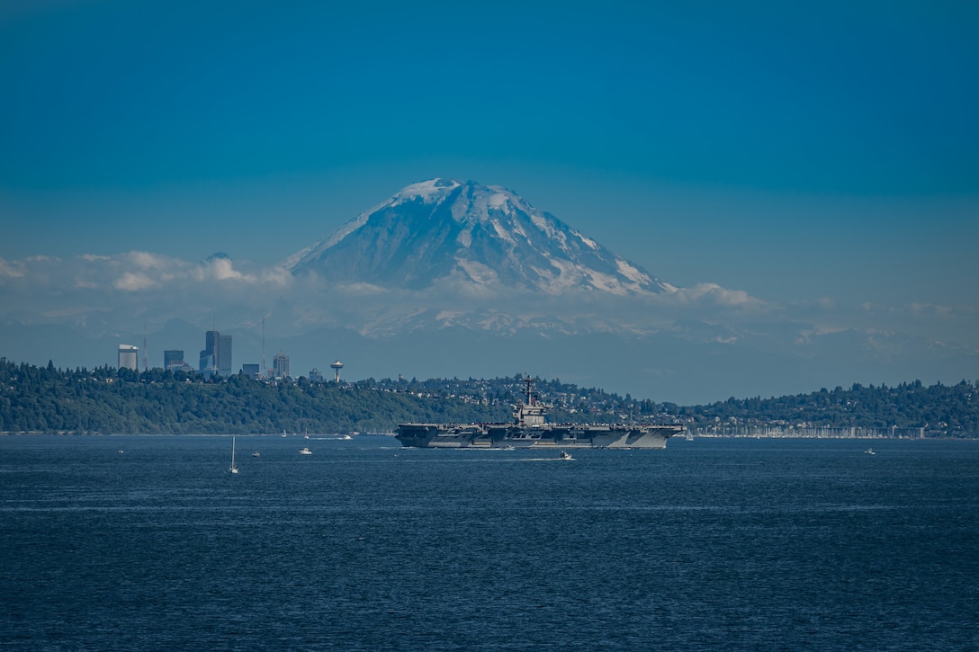 An aircraft carrier moves through the water near the shoreline with a huge mountain in the background.