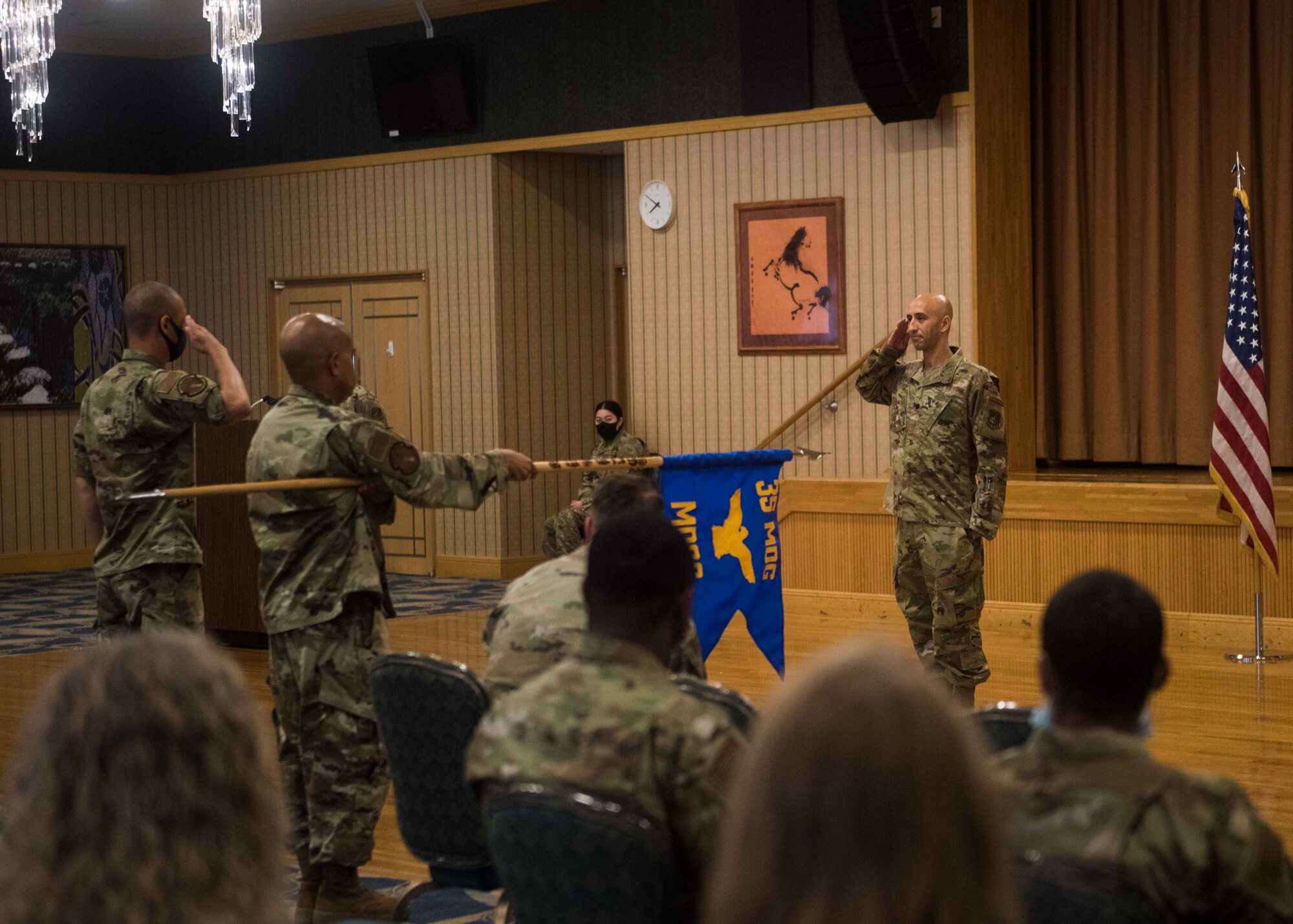 A service member on the right renders a salute to another service member and a guidon bearer while audience members watch.