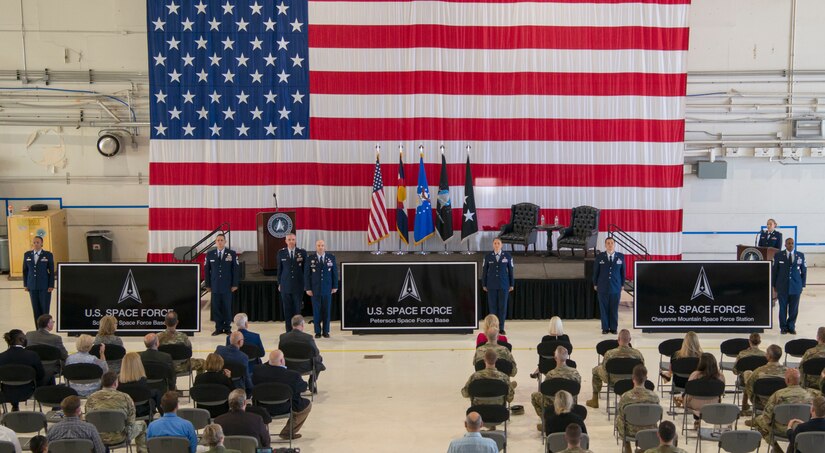 A crowd of people watch military leaders perform a ceremony in front of a large American flag.