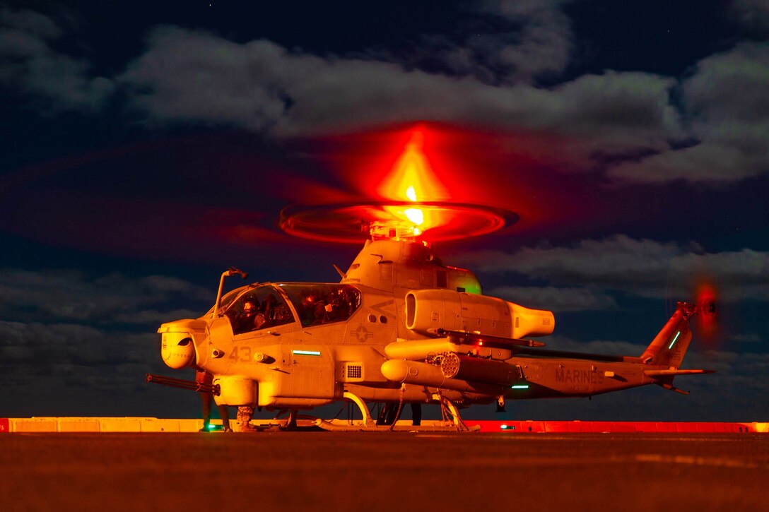 A helicopter in yellow light sits on the flight deck of a ship at night with rotors moving.