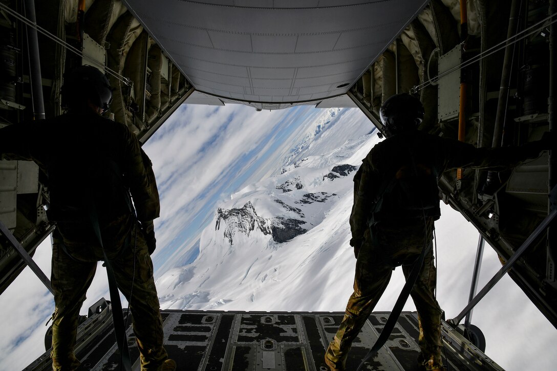 Two airmen look out over snowy mountains from an open aircraft door.