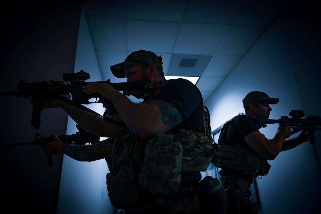 Three airmen hold weapons as they move through a building illuminated by blue light.