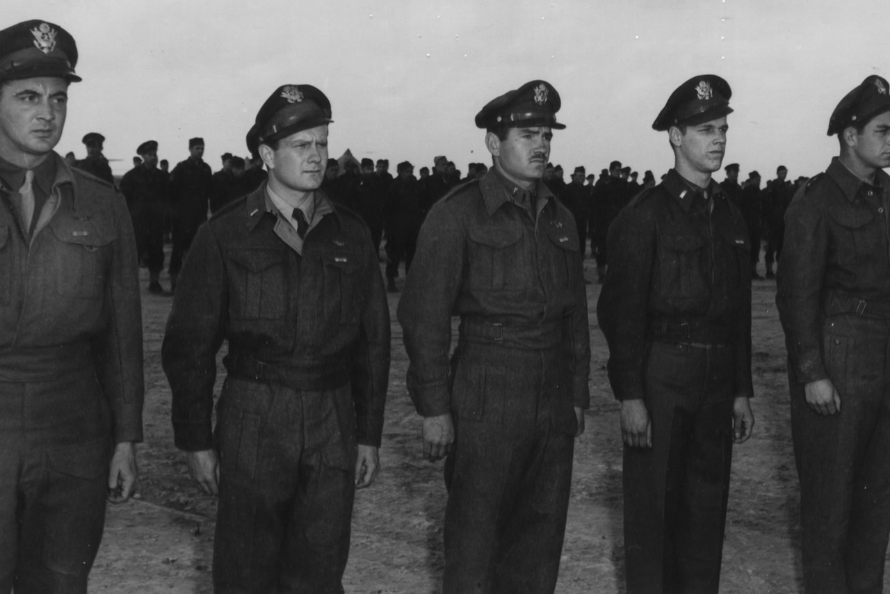 Five men in uniform stand side by side at attention. Dozens of other men stand at attention behind them.