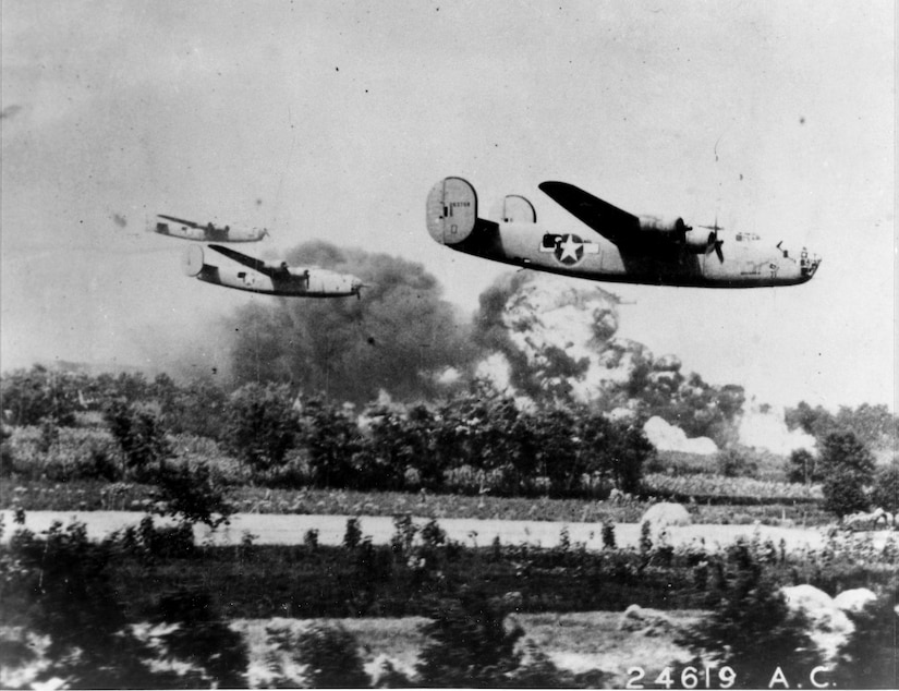 Three bomber aircraft fly low over land. Large plumes of smoke and fire are visible in the background.