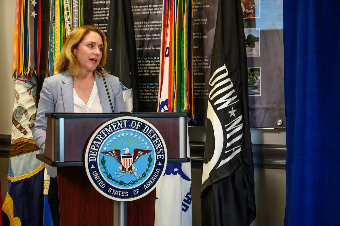 A woman stands behind a lectern.  Behind her is a flag with the POW/MIA logo.