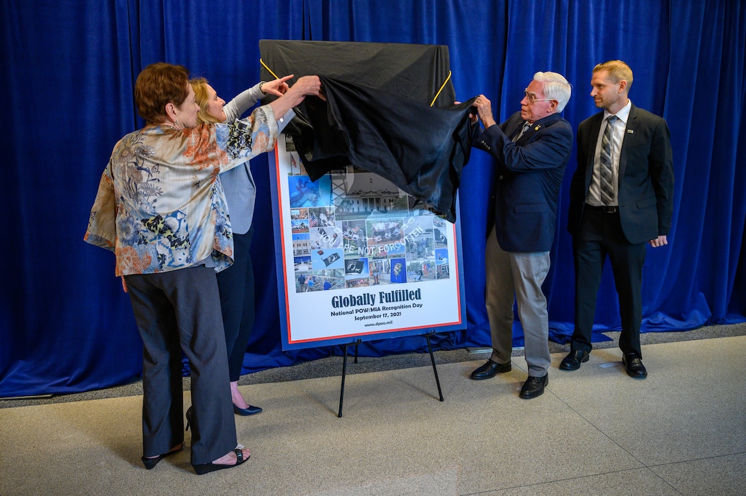 Four people lift a drape off a poster, which is sitting on an easel.