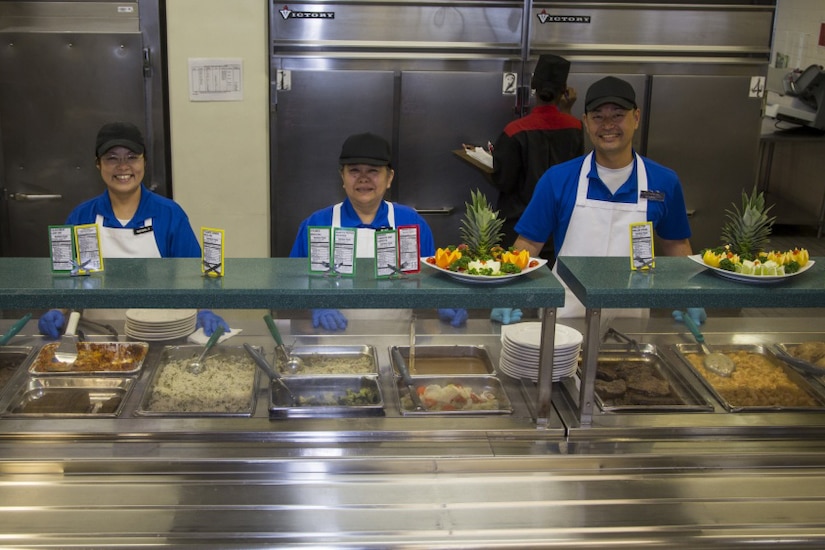 Three people stand behind the serving line at a cafeteria.