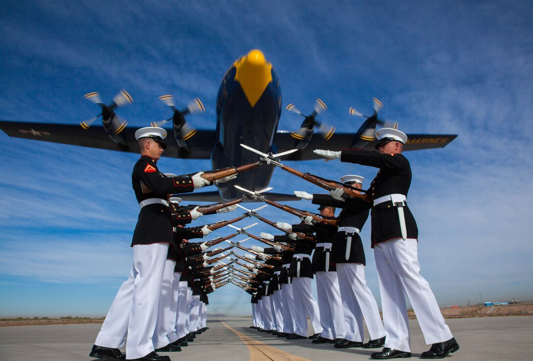 A group of Marine do rifle drills on a flightline as a blue aircraft flies low overhead.