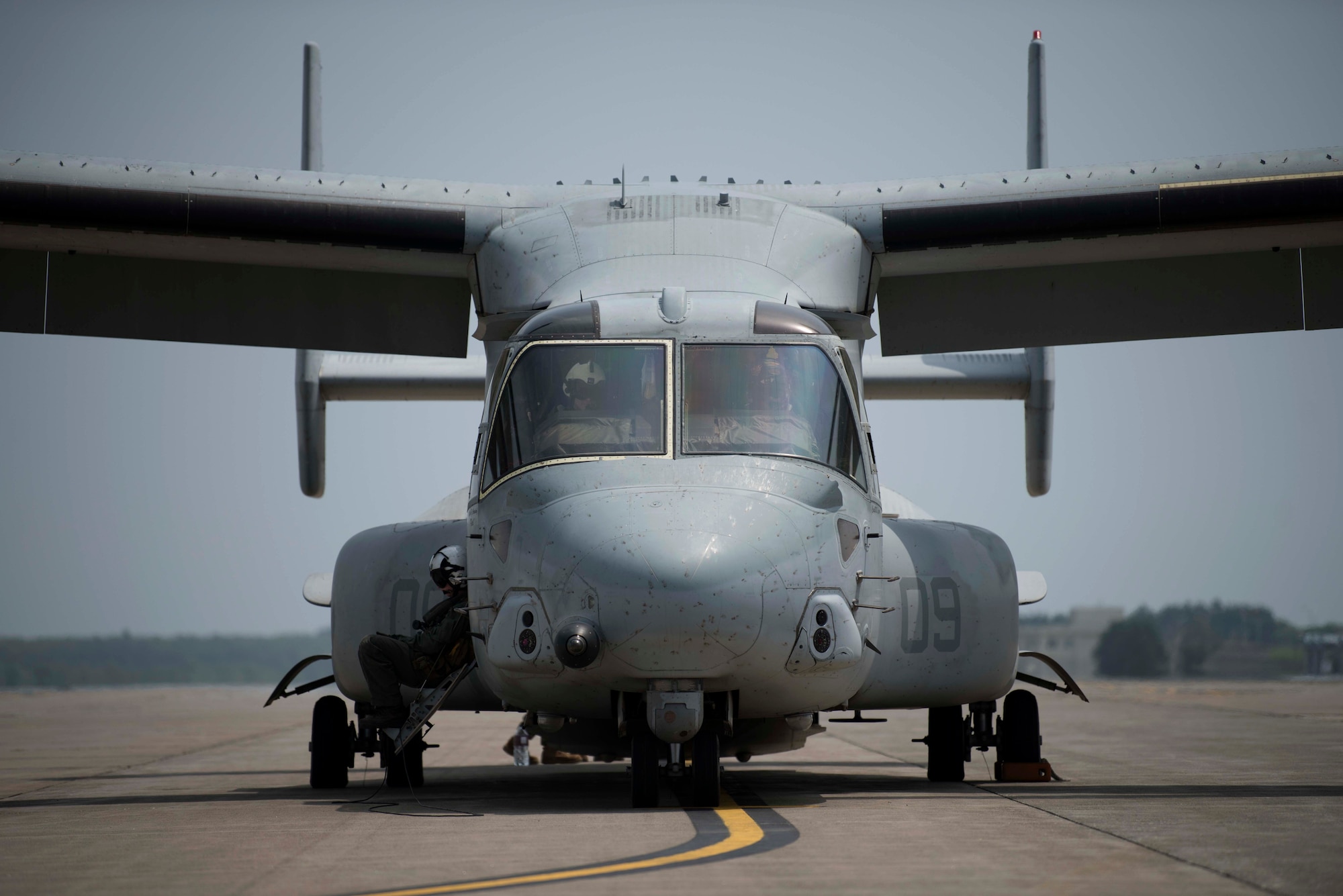 A grey aircraft sits on a flight line while individuals in flight suits operate it