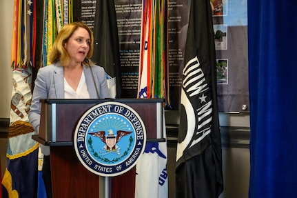 A woman stands behind a lectern.  Behind her is a flag with the POW/MIA logo.