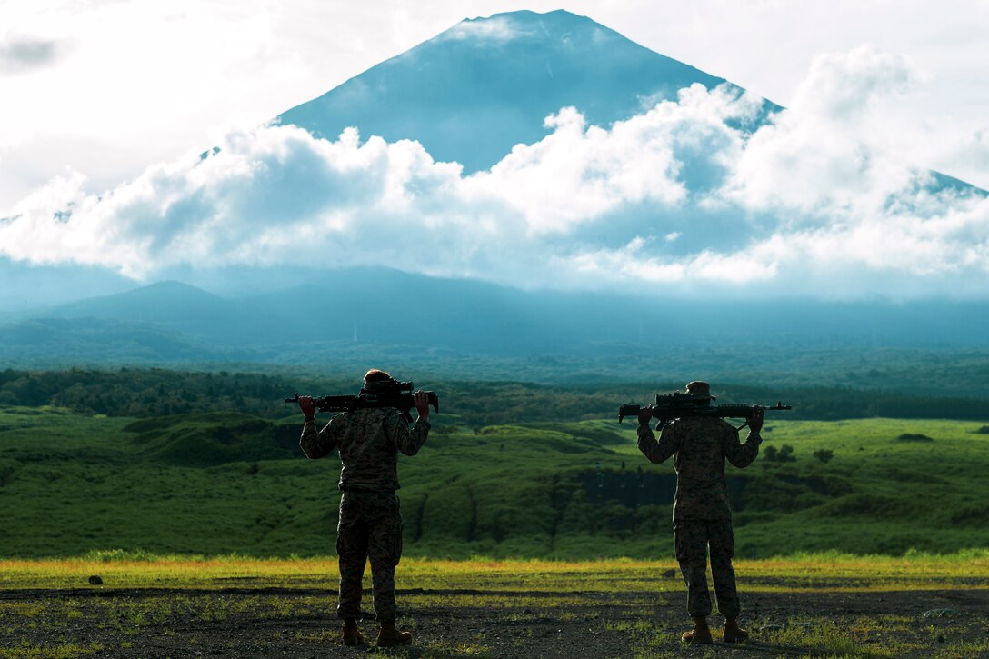 Two Marines, shown from behind, stare at a mountain while holding rifles.
