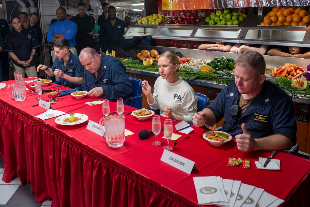Sailors seated at a table taste dishes while others observe.