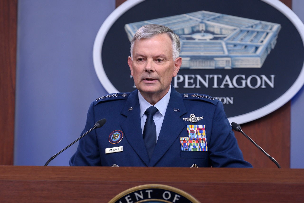 A  man wearing a military uniform stands at a lectern and appears to speak into two microphones.
