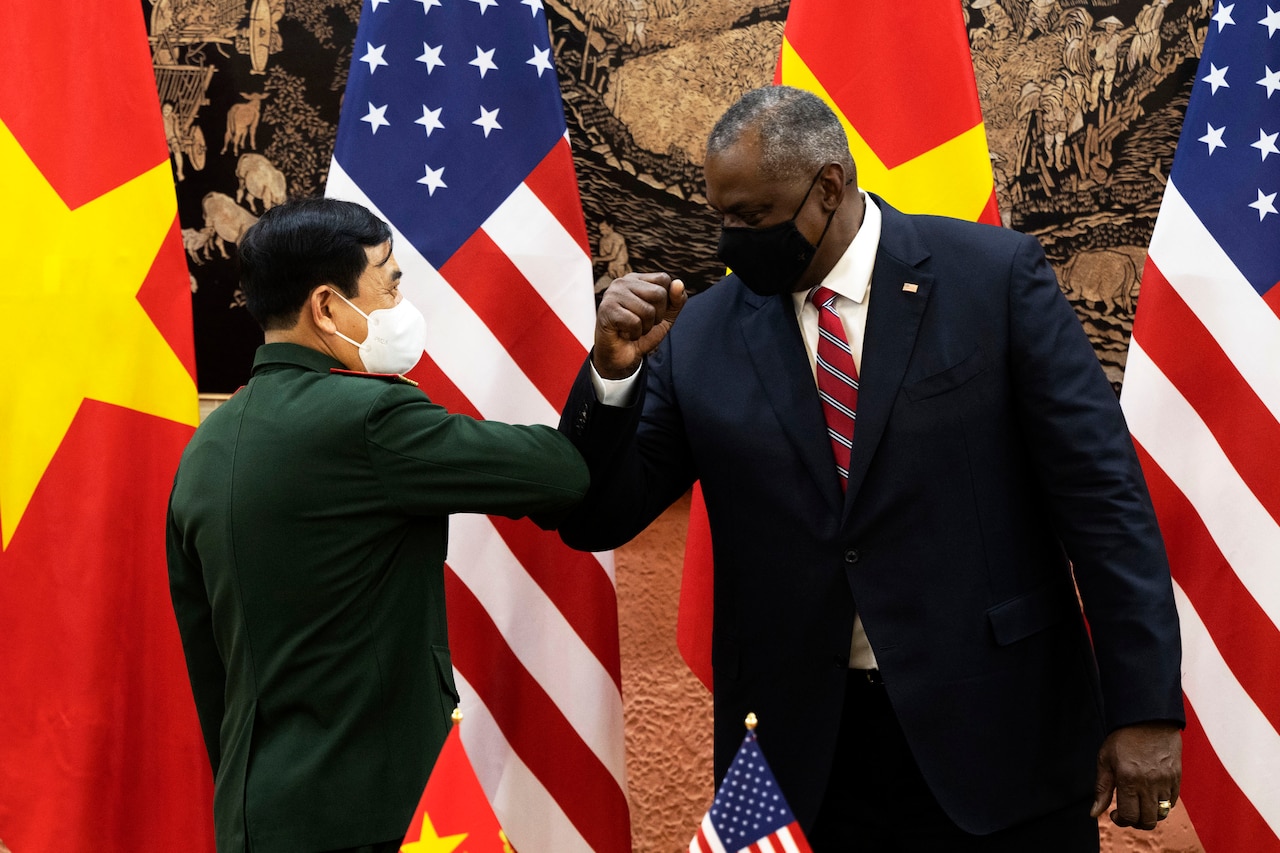 The U.S. Defense Secretary and Vietnamese Defense Minister bump elbows in front of country flags.
