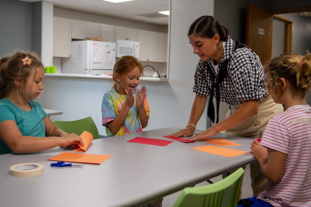 An airman in civilian clothes speaks to three children sitting around a table.