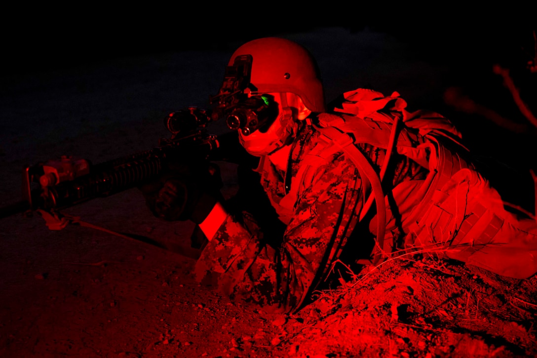 A Marine illuminated by red light looks through the scope of a weapon.