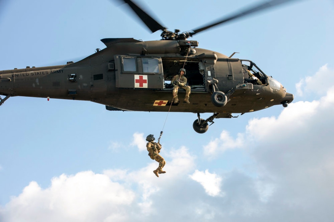 A soldier holds on to a rope attached to an airborne helicopter as a fellow soldier watches.