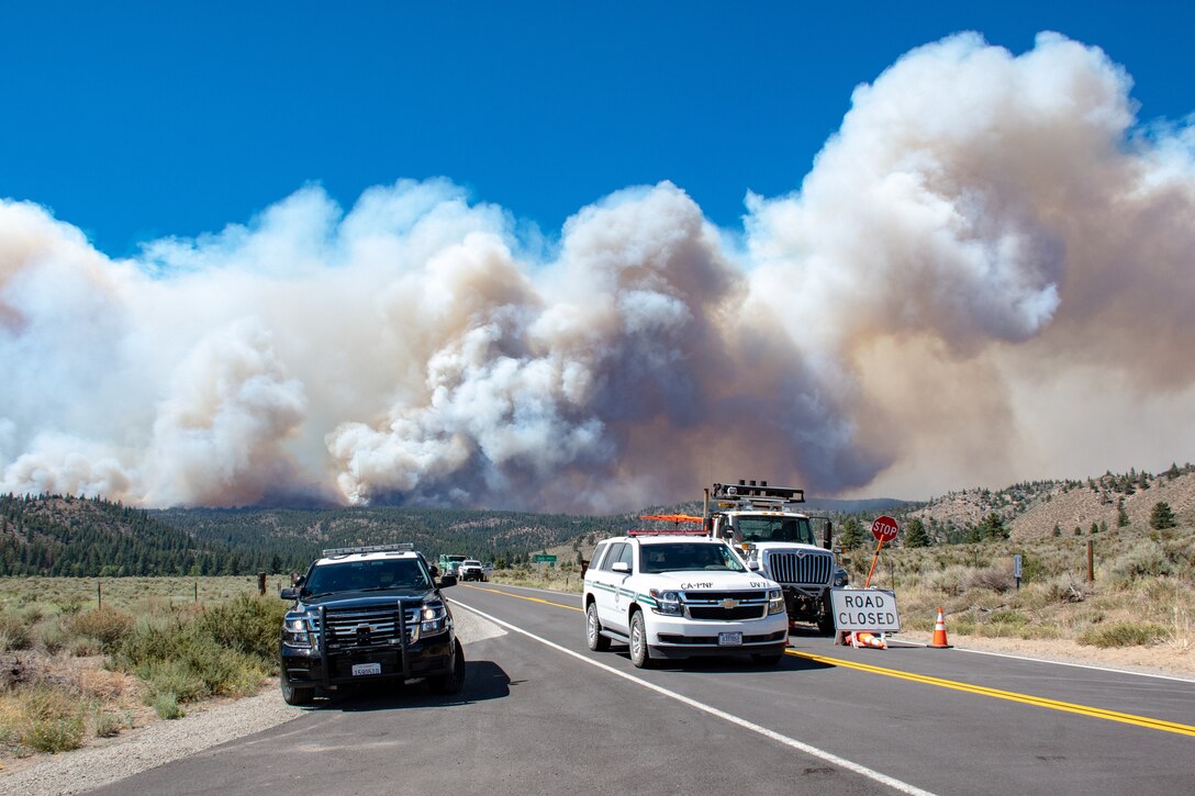 Official vehicles block a highway. In the background, there are clouds of smoke.