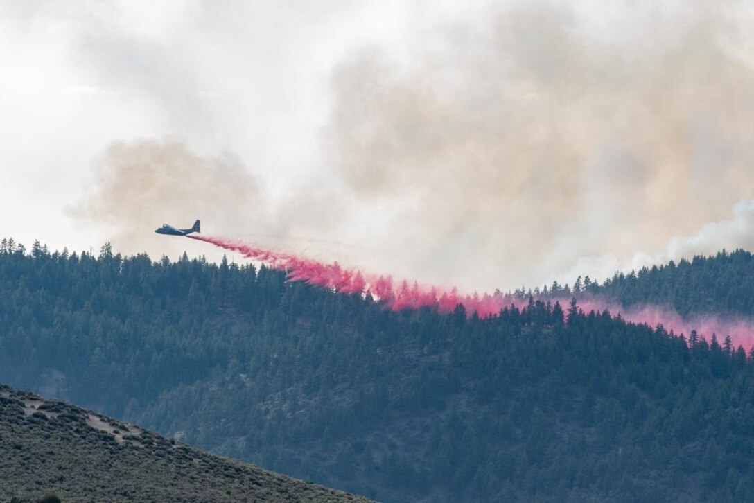 An aircraft moves across a green landscape, leaving behind a trail of red fire retardant.