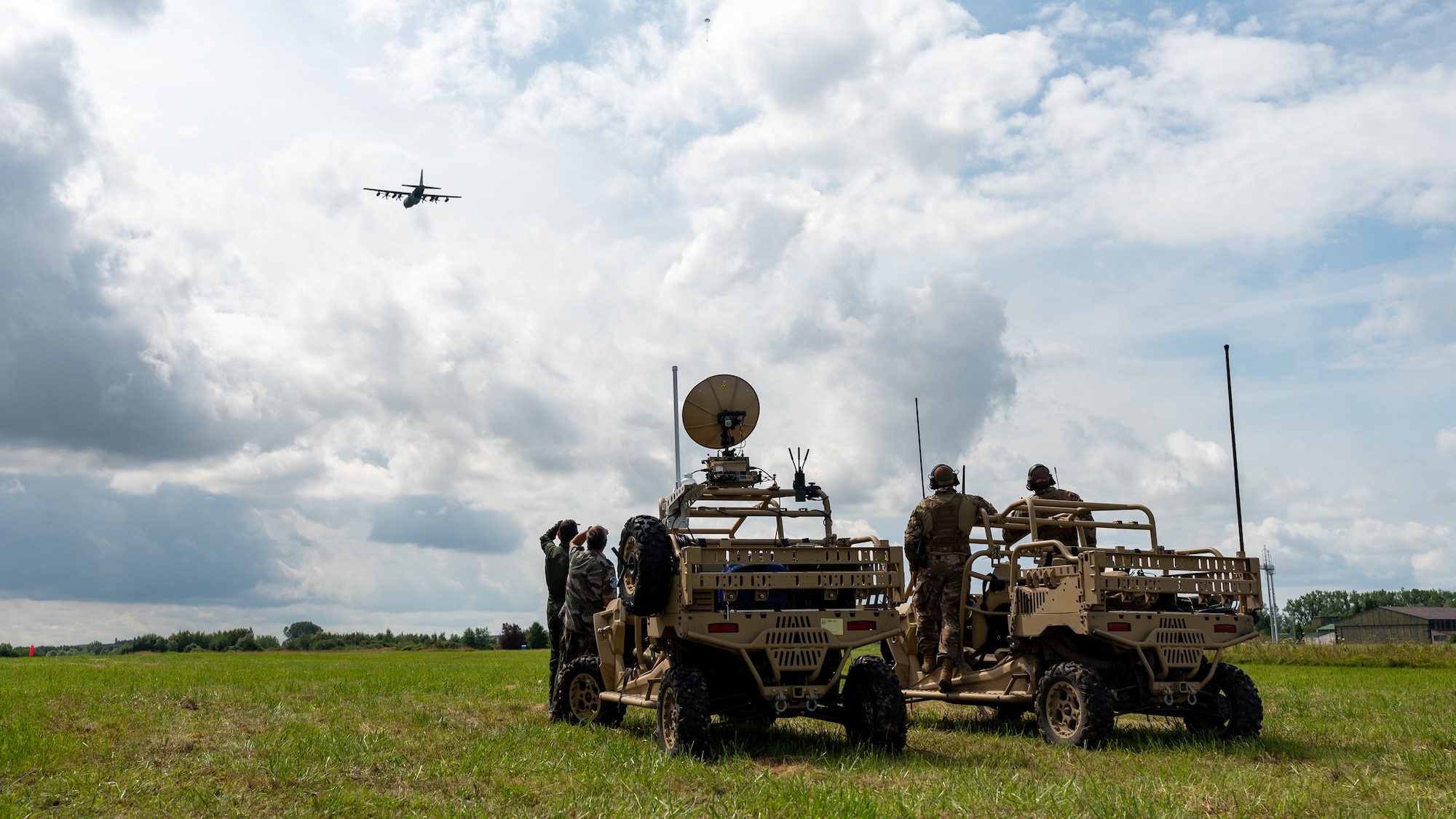People standing next to vehicles in a field and looking at an aircraft flying in the distance.